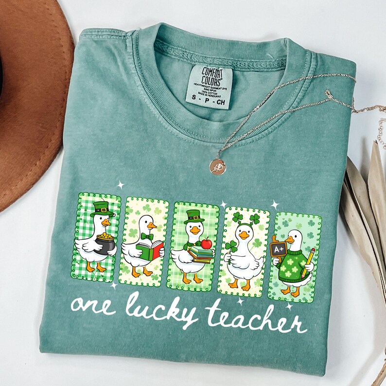 May include: A seafoam green t-shirt featuring a graphic of five cartoon ducks in St. Patrick's Day attire. Each duck is in a framed design with a green and white checkered background. The text "one lucky teacher" is printed below the ducks.