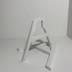 May include: White plastic easel stand with a simple design. The stand is made of two pieces that are connected together. The stand is designed to hold a small object, such as a picture frame or a small sculpture.
