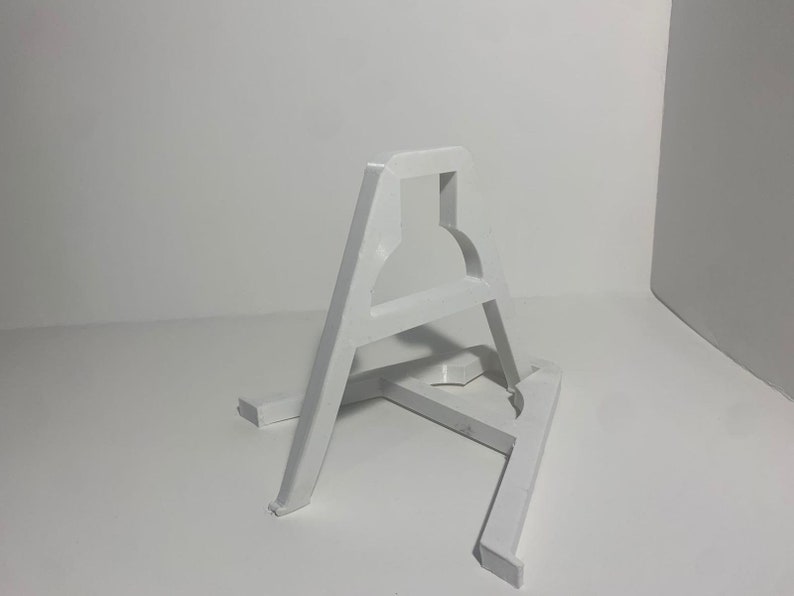 May include: White plastic easel stand with a simple design. The stand is made of two pieces that are connected together. The stand is designed to hold a small object, such as a picture frame or a small sculpture.