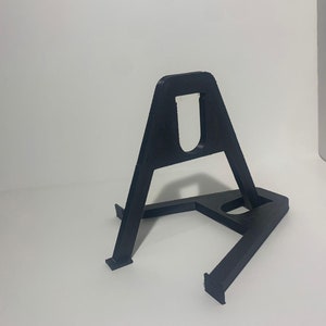 May include: A black plastic stand with a triangular base and a U-shaped cutout in the top. The stand is designed to hold a small object, such as a phone or a tablet.