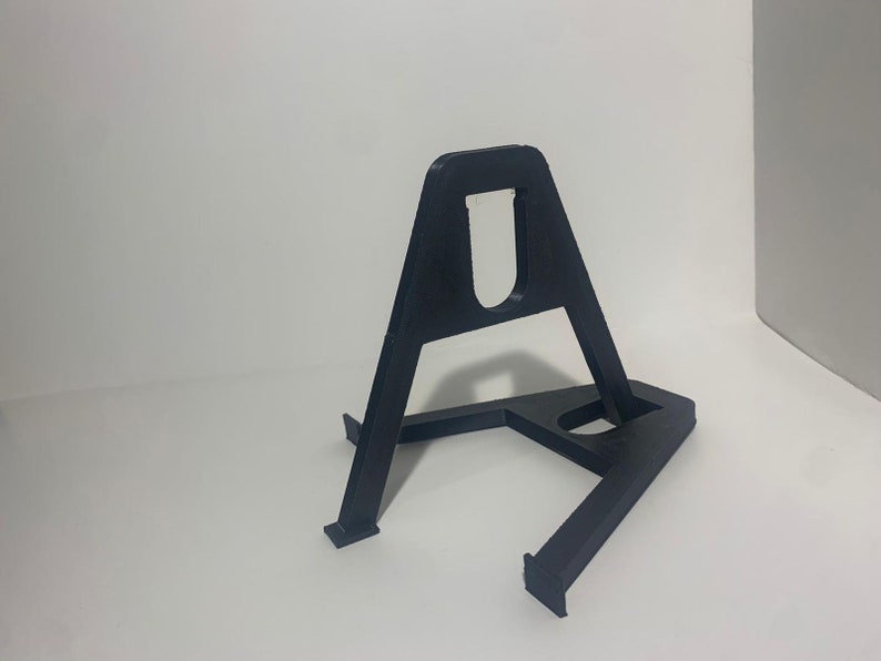 May include: A black plastic stand with a triangular base and a U-shaped cutout in the top. The stand is designed to hold a small object, such as a phone or a tablet.