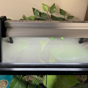 May include: A black metal reptile heat lamp fixture with a silver heat lamp. The lamp is mounted on a black metal stand with two legs. The lamp is on top of a black mesh terrarium.