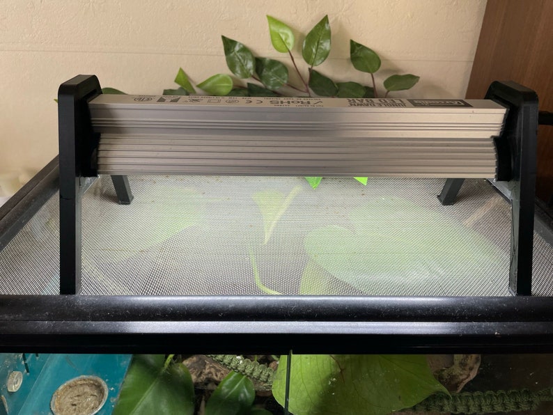 May include: A black metal reptile heat lamp fixture with a silver heat lamp. The lamp is mounted on a black metal stand with two legs. The lamp is on top of a black mesh terrarium.