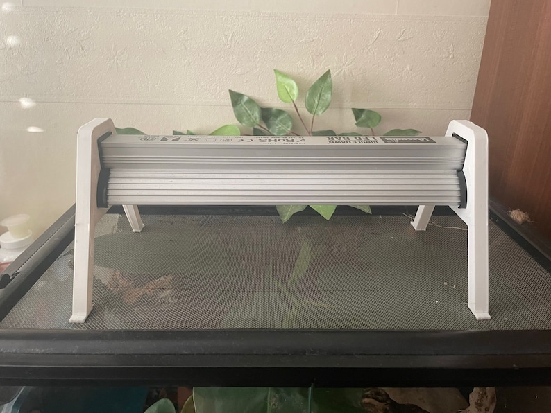 May include: White plastic stand with a white light fixture on top. The light fixture has a black trim and the text "Reptile UVB 10.0" printed on it.