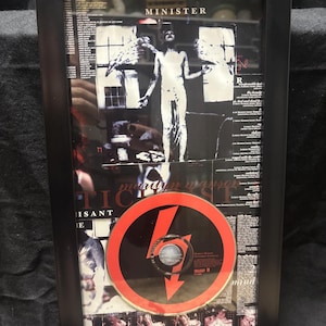 May include: Framed artwork featuring a CD with a red and black design, a black and white image of a person with wings, and the word "MINISTER". The frame is black.