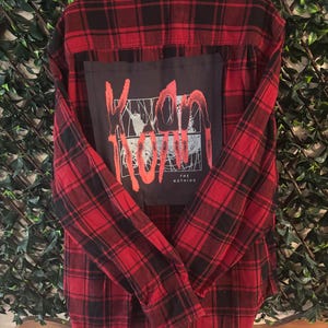 May include: Red and black plaid flannel shirt with a black patch on the back. The patch features the band name "Korn" in red lettering, along with the words "The Nothing". The shirt is displayed against a green leafy background.