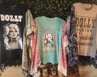 May include: Three t-shirts featuring Dolly Parton graphics. The left is black with a portrait and the word DOLLY. The center is light green with a portrait and DOLLY PARTON LIVE IN CONCERT text. The right is tan with DOLLY PARTON LIVE IN CONCERT.