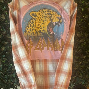 May include: A long-sleeved, button-down shirt with a plaid pattern in shades of orange and white. The back features a pink patch with a roaring leopard graphic and the band name "Def Leppard" in gold lettering.