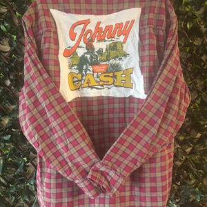 May include: Red and brown plaid button-down shirt with a white patch on the back. The patch features the text "Johnny Cash" with an illustration of a cowboy on a horse in a desert landscape. The shirt has long sleeves.