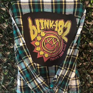 May include: Green, black, yellow, and white plaid flannel shirt with a large black patch on the back. The patch features the band name "BLINK-182" in yellow and a red and yellow graphic with the words "CRAPPY PUNK ROCK."
