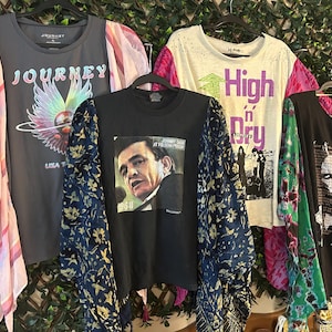 May include: Four graphic t-shirts hanging on a rack. One is gray with "JOURNEY" and a colorful graphic. Another is black with a Johnny Cash image. A third is white with "High n' Dry" text. The last is black with a face graphic.