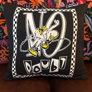 May include: Black square pillow with a white and black checkered border. The pillow features the words "NO DOUBT" in a stylized font, with a cartoon figure holding a microphone. The figure is yellow and white.