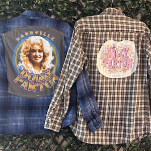 May include: Two long-sleeve button-down shirts. The left shirt is blue plaid with a gray back panel featuring a portrait of Dolly Parton and the text Nashville Dolly Parton. The right shirt is brown plaid with a white patch featuring pink text Dolly Parton.