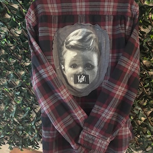 May include: A red, black, and gray plaid flannel shirt with a black and white graphic on the back. The graphic is a portrait of a child with the word "Korn" on a small black tag.