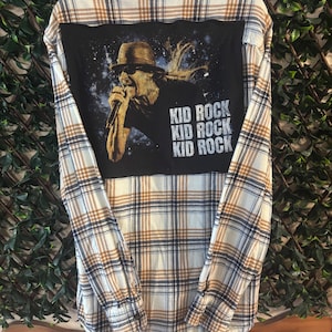 May include: A long-sleeved flannel shirt with a plaid pattern in cream, brown, and navy blue. The back features a black panel with a graphic of Kid Rock and the text "KID ROCK" repeated three times. The shirt is displayed on a green leafy background.