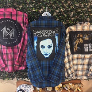 May include: Three plaid flannel shirts with band patches. The pink shirt has a black patch with the text "Sleep Token". The blue shirt features an "Evanescence Fallen" patch. The third shirt is tan and white with a "Kid Rock" patch.