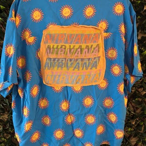 May include: A short-sleeved, button-down shirt in a vibrant blue, adorned with repeating yellow sun graphics. The back features a large, orange patch with the word "NIRVANA" printed in a layered, colorful design. The shirt is made of a lightweight fabric.