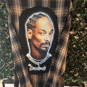 May include: A black and brown plaid flannel shirt with a graphic print of Snoop Dogg on the back. The graphic includes a chain necklace and the text "Dogg Supply".