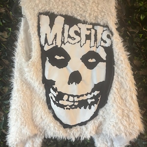 May include: A white, fluffy vest featuring a large black and white graphic of a skull with the word "MISFITS" above it. The vest has a soft, textured appearance and a sleeveless design. The graphic is centered on the front of the vest.