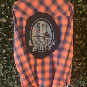 May include: Orange and navy blue plaid button-down shirt with a black patch on the back. The patch features a graphic with the text "Flogging Molly". The shirt has long sleeves and a classic collar.