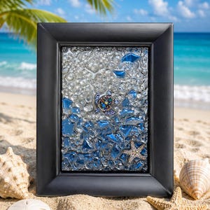 Puede incluir: Obra de arte enmarcada con un mosaico de vidrio transparente y azul, con una tortuga marina enjoyada. El marco negro descansa sobre arena con conchas marinas y estrellas de mar. El fondo muestra una playa y un océano.