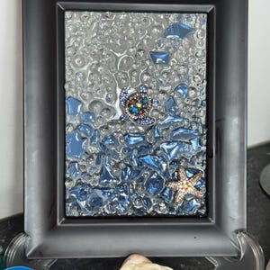 Può includere: Arte a mosaico incorniciato con una tartaruga marina e una stella marina. L'opera d'arte utilizza pezzi di vetro trasparente e blu per raffigurare una scena oceanica. La cornice nera circonda il mosaico, e la tartaruga e la stella marina sono impreziosite da gemme colorate.