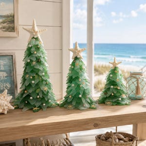 May include: Three decorative green glass Christmas trees with starfish toppers, pearl and starfish accents. The trees are arranged on a wooden table with a coastal theme, near a window overlooking the ocean.