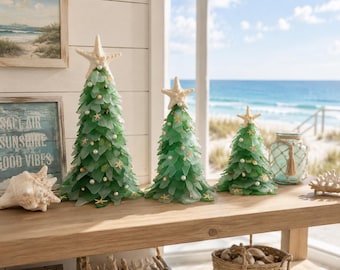May include: Three decorative green glass Christmas trees with starfish toppers, pearl and starfish accents. The trees are arranged on a wooden table with a coastal theme, near a window overlooking the ocean.