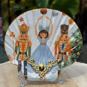 May include: A decorative seashell featuring a painted scene of a ballerina and two nutcracker figures. The ballerina wears a blue dress and bow. The shell is embellished with a gold-colored decorative element and is displayed on a clear stand.