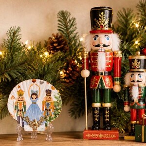 May include: A decorative holiday display featuring a large wooden nutcracker soldier in a red and green uniform, a smaller nutcracker, and a decorative plate with a ballerina and nutcracker figures. The background includes pine branches and fairy lights.