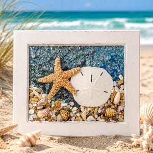 May include: A framed beach scene with a white wooden frame. Inside, a starfish and sand dollar rest on a bed of small shells and pebbles against a blue, textured background. The frame sits on sand with the ocean in the background.
