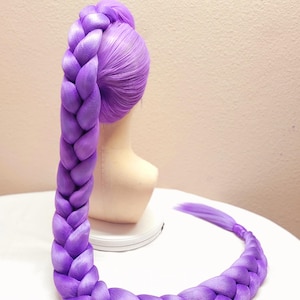 May include: A 50-inch dark purple wig styled in a long, thick, single braid that extends from the crown of the head down to the base. The wig features a smooth, vibrant purple fiber texture and includes tulle detailing at the top attachment point.
