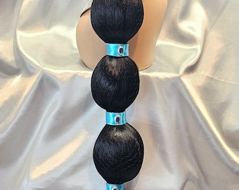 Può includere: Una lunga parrucca nera con un chignon in cima e una coda di cavallo con nastri blu e gioielli. La parrucca è su una testa di manichino.