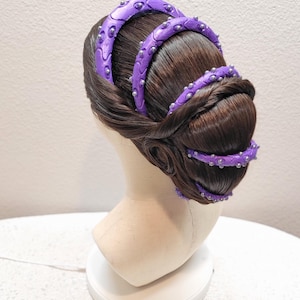 May include: A dark brown wig styled in a low bun with three purple, curved, textured headbands featuring small silver studs. The headbands are arranged in a tiered, parallel fashion across the top and back of the hair, creating a structured look.