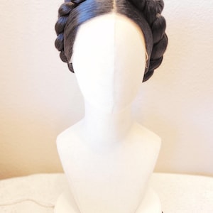May include: A dark brown wig styled with a thick, braided crown that encircles the head. The hair is parted in the center and pulled back into a smooth, rounded braid that sits high on the head, displayed on a plain white mannequin head.