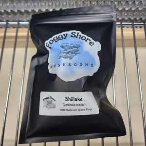 May include: A black resealable bag with a white logo that says "Foggy Shore Mushrooms" and a mushroom illustration. The bag contains 100 mushroom spawn plugs and is labeled "Shiitake (Lentinula edodes)"