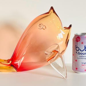 May include: A large, hand-blown glass sculpture of a fish with a red and orange ombre finish. The fish has a long, pointed snout and a tail that curls up. The sculpture is sitting on a white surface next to a can of Bubbly Bounce caffeinated sparkling water.