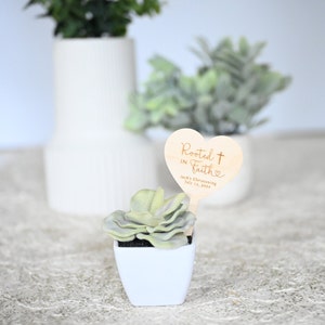 K&ouml;nnte beinhalten: Eine kleine Sukkulente in einem wei&szlig;en quadratischen Topf mit einem herzf&ouml;rmigen Schild aus Holz, auf dem "Rooted in Faith" mit einem Kreuz und einem Herzen steht. Das Schild enth&auml;lt auch den Text "Jack's Christening July 10, 2022".