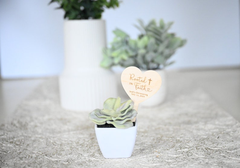 K&ouml;nnte beinhalten: Eine kleine Sukkulente in einem wei&szlig;en quadratischen Topf mit einem herzf&ouml;rmigen Schild aus Holz, auf dem "Rooted in Faith" mit einem Kreuz und einem Herzen steht. Das Schild enth&auml;lt auch den Text "Jack's Christening July 10, 2022".