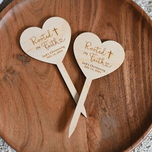 K&ouml;nnte beinhalten: Zwei herzf&ouml;rmige Schilder aus Holz mit dem Text "Rooted in Faith" und einem Kreuz. Die Schilder sind auf Holzst&auml;bchen und eignen sich perfekt f&uuml;r eine Taufe.