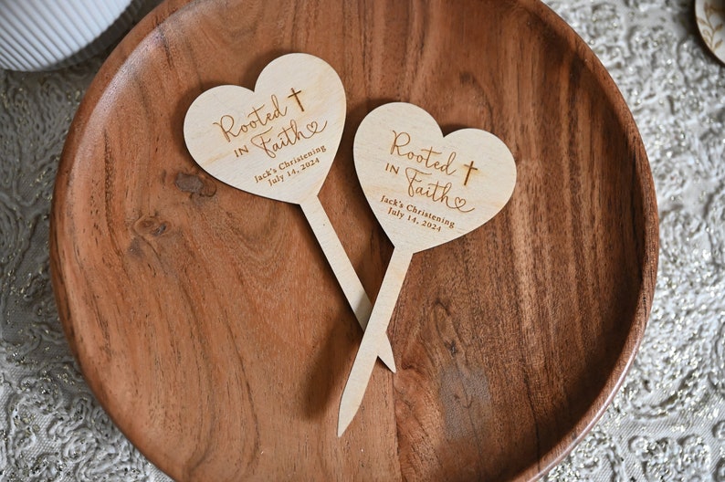 K&ouml;nnte beinhalten: Zwei herzf&ouml;rmige Schilder aus Holz mit dem Text "Rooted in Faith" und einem Kreuz. Die Schilder sind auf Holzst&auml;bchen und eignen sich perfekt f&uuml;r eine Taufe.