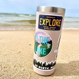 May include: A stainless steel tumbler with a beige exterior and multiple stickers. The stickers feature a blue and white "To the Lake" design, a black and white "Up North, Michigan" design, and a blue and yellow "Michigan Explore" sticker with the text "Water - Winter Wonderland".