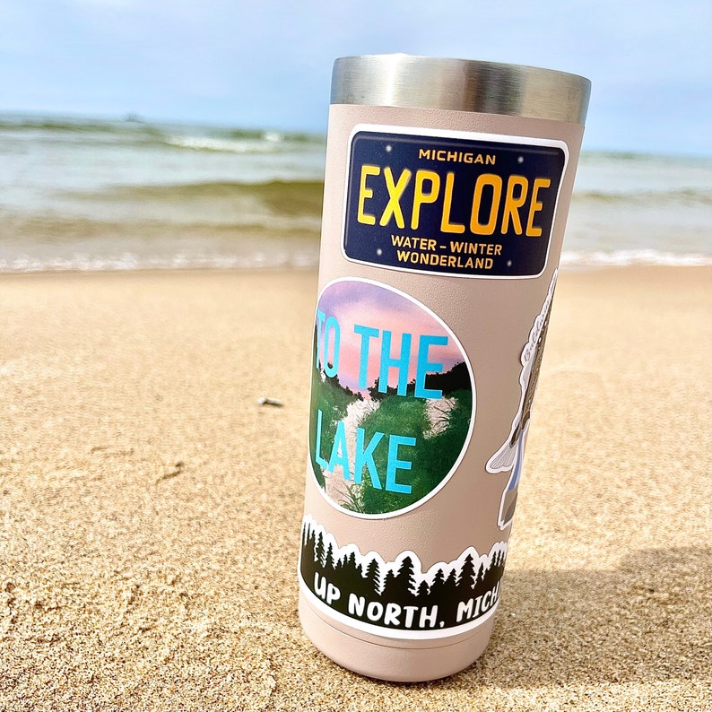 May include: A stainless steel tumbler with a beige exterior and multiple stickers. The stickers feature a blue and white "To the Lake" design, a black and white "Up North, Michigan" design, and a blue and yellow "Michigan Explore" sticker with the text "Water - Winter Wonderland".