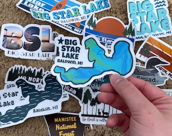 May include: A collection of stickers featuring the text "Big Star Lake" and "Baldwin, MI". The stickers are in various colors and designs, including a blue and green lake, a brown and orange sunset, and a black and white forest scene. The stickers are arranged on a sandy surface.