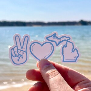 May include: Three blue stickers on a hand. The stickers are a peace sign, a heart, and the outline of the state of Michigan.