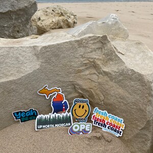 May include: Assortment of colorful stickers featuring Michigan-themed designs, including a smiley face with the word "Michigan" and the phrase "fresh coast" repeated four times. Other stickers include a map of Michigan, a forest scene with the text "UP NORTH. MICHIGAN", and a rainbow-colored sticker with the word "OPE".