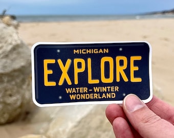 May include: A blue and yellow Michigan license plate sticker with the word "EXPLORE" in large yellow letters. The sticker also says "WATER - WINTER WONDERLAND" in smaller yellow letters.