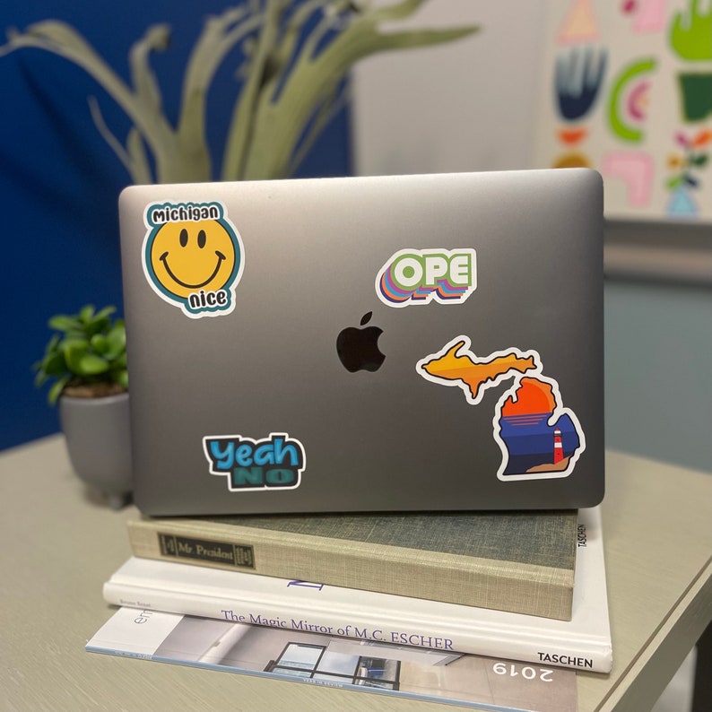 May include: A silver laptop computer with several stickers on the lid. The stickers include a smiley face with the text "Michigan nice", a state outline of Michigan with a sunset scene, the word "OPE" in a rainbow font, and the words "Yeah No" in a blue and green font.