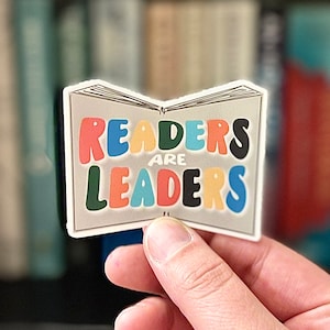 Könnte beinhalten: Ein Aufkleber in Form eines offenen Buches mit dem Text "READERS ARE LEADERS" in bunten Buchstaben. Der Aufkleber wird von einer Hand gehalten, mit einem verschwommenen Hintergrund von Bücherregalen.