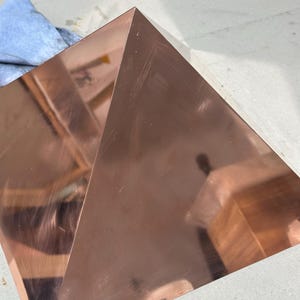 Könnte beinhalten: Eine Kupferpyramide mit einer glatten, polierten Oberfläche. Die Pyramide steht auf einer hellgrauen Oberfläche.
